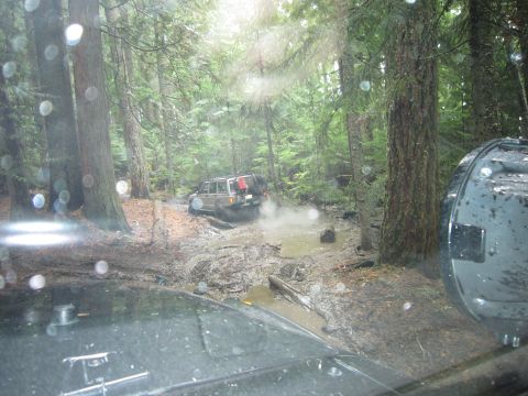 2008 Jeep Wrangler - A Jeep Cherokee XJ navigates a muddy trail in a forest, with another vehicle's perspective visible in the foreground.