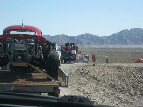 1986 Ford Bronco - A convoy of vehicles travels down a dusty desert road, with a custom dune buggy being towed on a trailer.