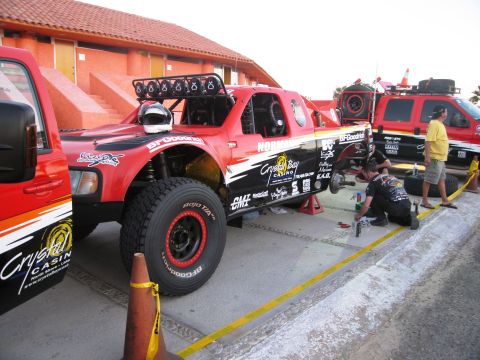 1986 Ford Bronco - A red off-road truck with numerous racing decals is shown being worked on, with another similar vehicle visible nearby.