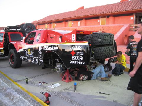 1986 Ford Bronco - Mechanics work on an off-road race truck, which has multiple sponsor decals and spare tires mounted.