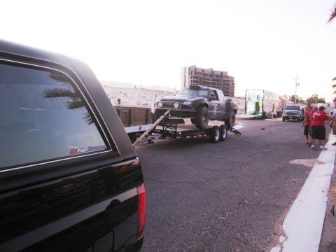 1986 Ford Bronco - A custom off-road pickup truck is loaded onto a trailer, being towed by another vehicle, with buildings and people in the background.