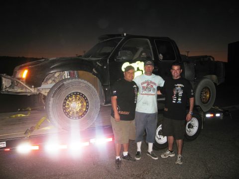 1986 Ford Bronco - Three men pose with a modified off-road truck loaded onto a trailer at night.