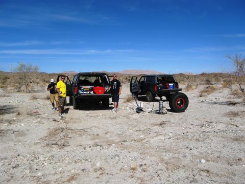 1986 Ford Bronco - Two Ford Broncos are parked in a desert setting, one is being worked on with a jack.