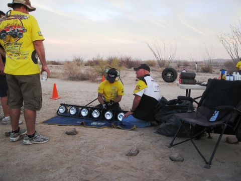 1986 Ford Bronco - A pit crew prepares equipment in a desert setting, with a spare tire and supplies nearby.