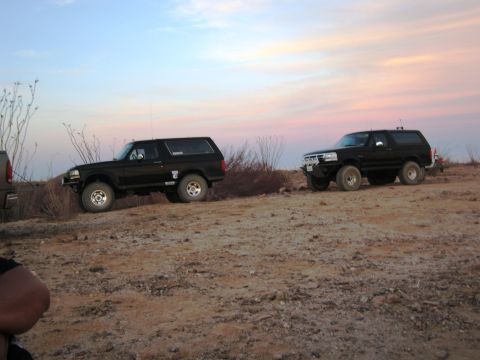 1986 Ford Bronco - Two black Ford Broncos are parked on a dusty hill during sunset, with a colorful sky overhead.