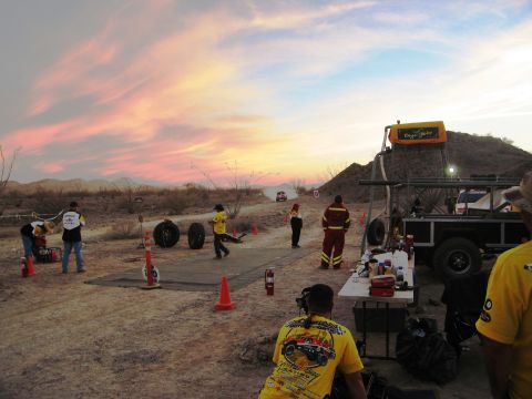 1986 Ford Bronco - A desert race pit stop is set up with people and vehicles under a colorful sunset sky.