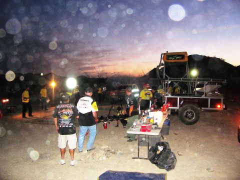 1986 Ford Bronco - A group of people are gathered around a trailer and table in a desert area at dusk, with lights on.