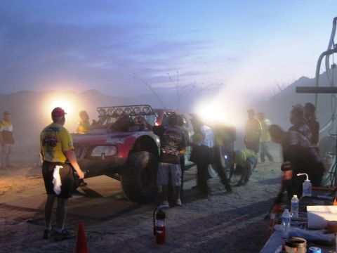 1986 Ford Bronco - A crew works on a red off-road race truck under bright lights as dusk settles over the desert landscape.