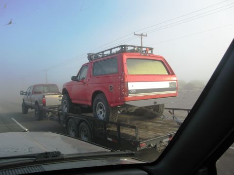 1986 Ford Bronco - A red Ford Bronco is transported on a trailer, with a pickup truck towing it through foggy conditions.