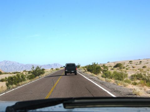 1986 Ford Bronco - A black SUV travels down a highway through a desert landscape under a clear blue sky.