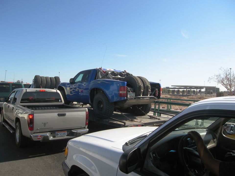 Blue off-road truck with flame decals is on a trailer behind a silver pickup. Another white truck is in the foreground