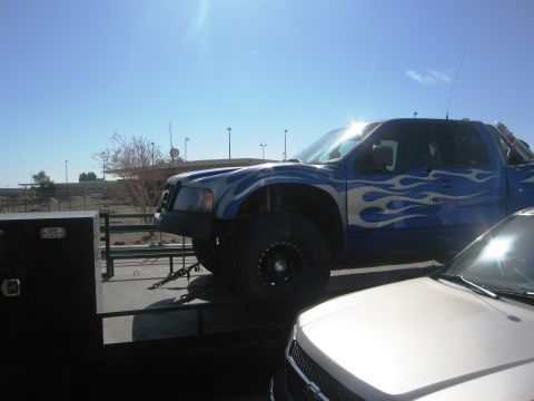 1986 Ford Bronco - A blue pickup truck with custom flame decals is loaded onto a flatbed trailer, with another vehicle partially visible in the foreground.