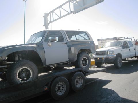 1986 Ford Bronco - A modified SUV is transported on a trailer, pulled by a pickup truck on a sunny day.