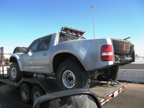 1986 Ford Bronco - A modified pickup truck is transported on a flatbed trailer under a clear blue sky.