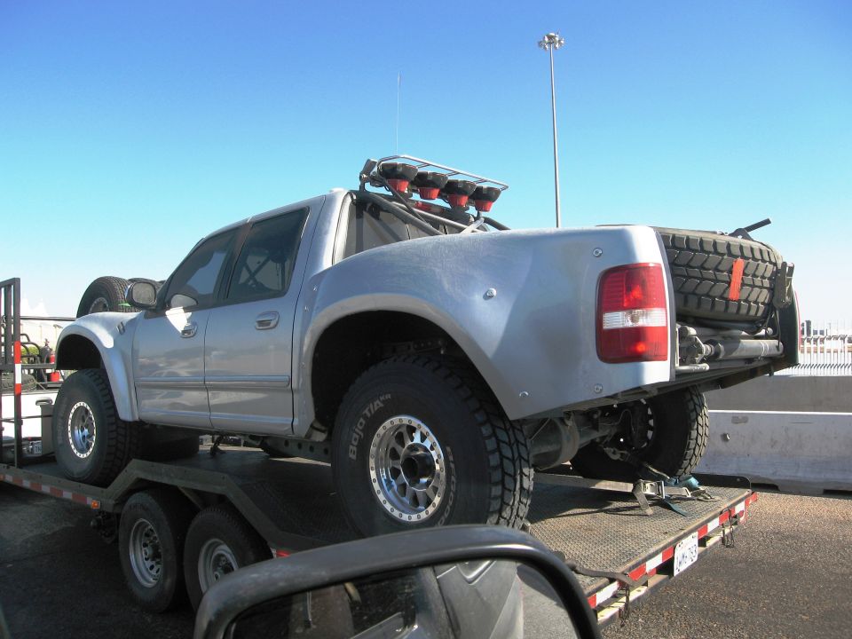 Silver pickup truck with off-road tires and light bar on a trailer, viewed from the rear quarter