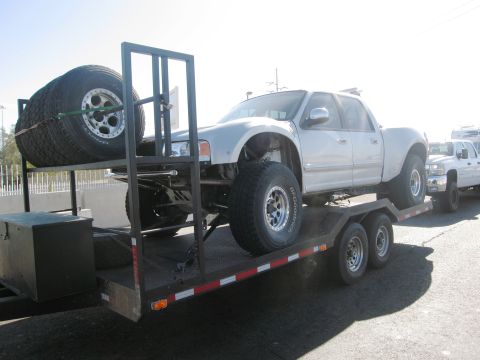 1986 Ford Bronco - A silver pickup truck, modified with large off-road tires, is transported on a trailer, with another truck following closely behind.