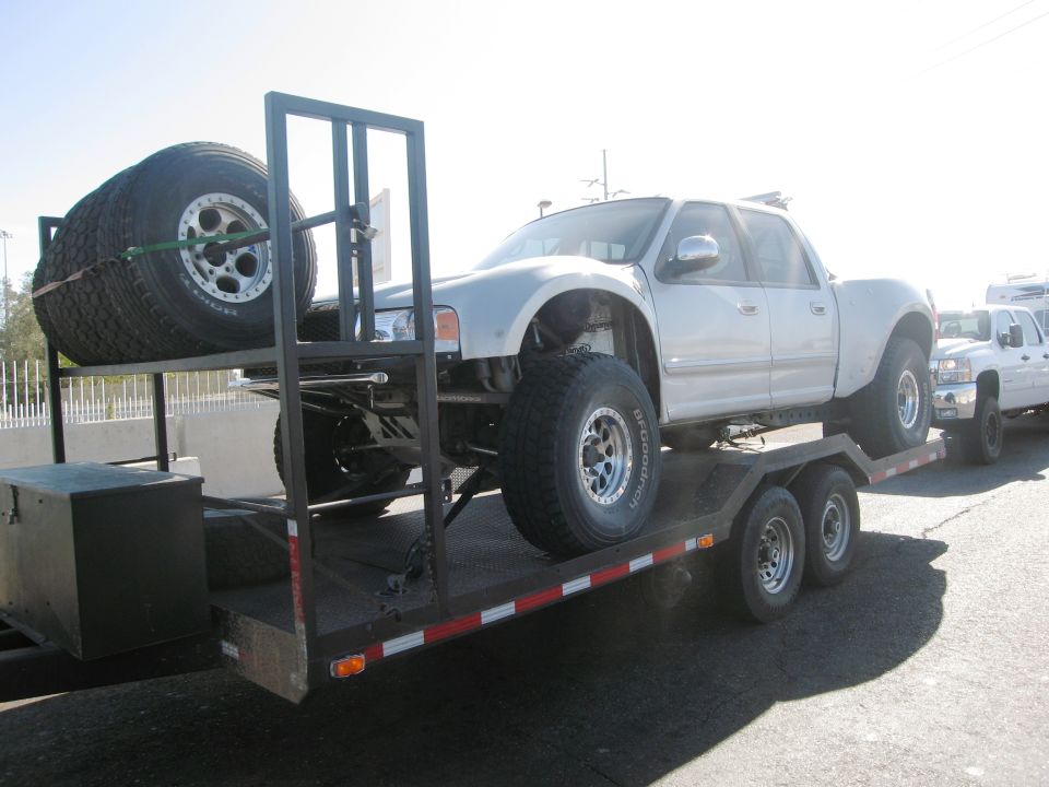 Silver pickup truck with oversized tires on a trailer, another truck visible behind