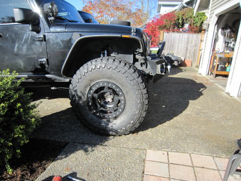 2008 Jeep Wrangler - Low front-quarter view of a lifted dark gray Jeep Wrangler in a residential driveway, focused on the driver's front wheel: a Toyo Open Coun…