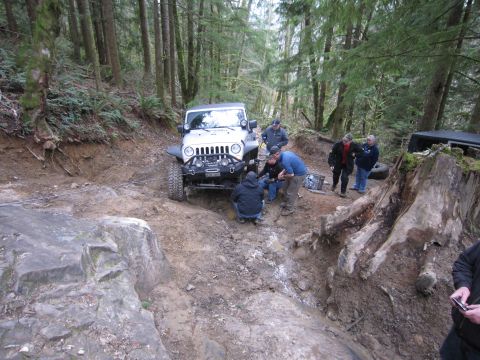 2008 Jeep Wrangler - A group of people work on a white 2008 Jeep Wrangler stuck on a muddy, wooded trail.