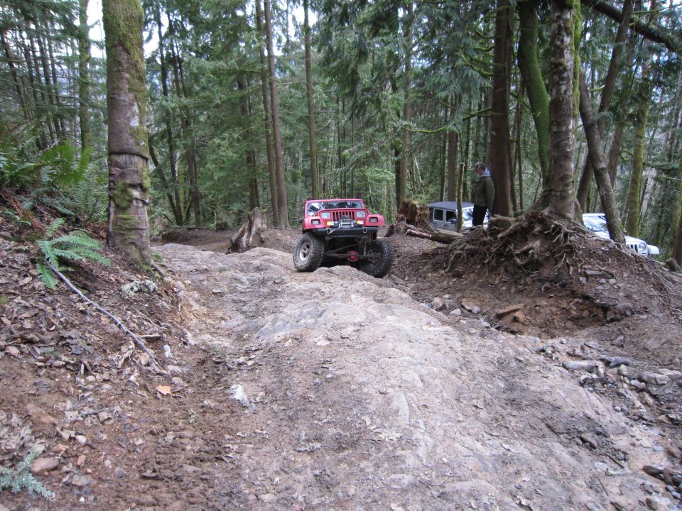 Red Jeep Wrangler driving up a rocky, wooded trail. Another Jeep and a white SUV are visible further up the path