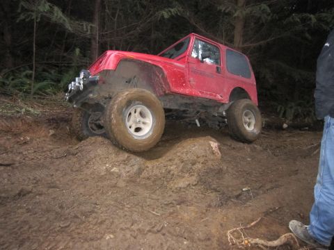 2008 Jeep Wrangler - Red Jeep Wrangler tackling a difficult muddy obstacle in a wooded area.