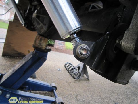 2008 Jeep Wrangler - Underside shot of a suspension job in a driveway, showing a new bare-aluminum shock body bolted through its lower eye to the axle bracket.…