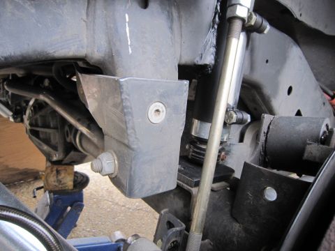 2008 Jeep Wrangler - Underbody close-up of a bare-metal fabricated bracket bolted to the frame rail beside a track bar and steering linkage, with a shock body a…