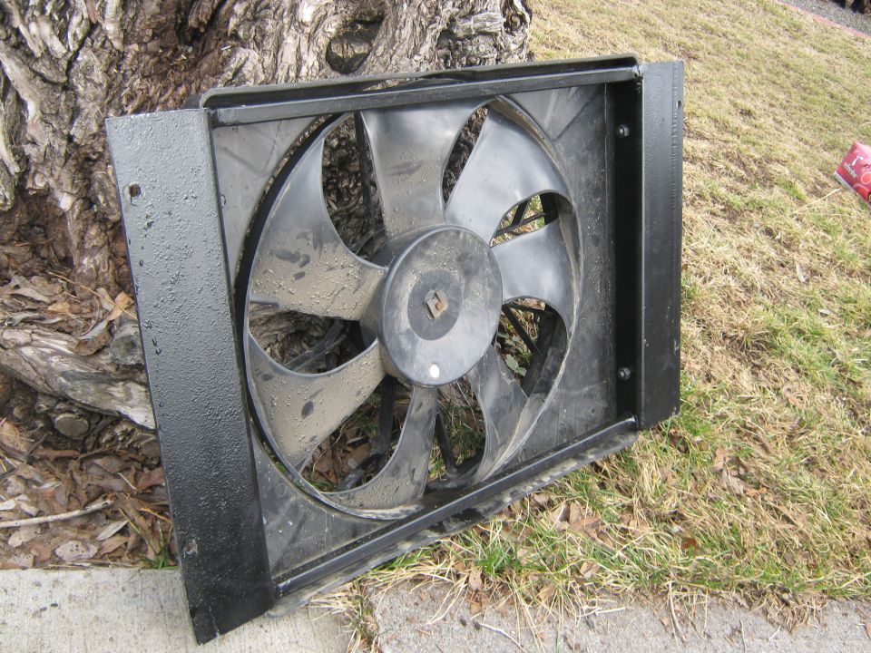 Radiator fan assembly with black metal frame and black plastic blades, resting on grass and dirt near a tree