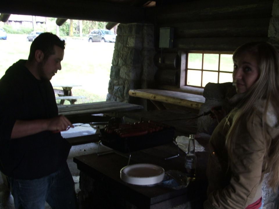 Two people grilling at a picnic shelter, with cars visible in the background