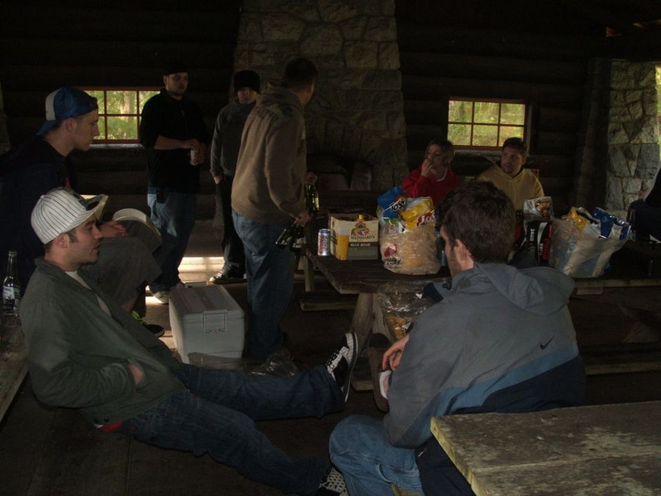 Group of people gathered in a wooden cabin with a stone fireplace, some sitting on benches and at a picnic table