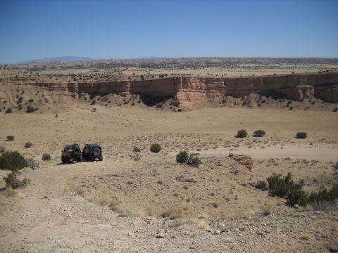 1986 Ford Bronco II - Two SUVs are parked on a dirt road in a vast desert landscape with a large canyon wall in the distance.