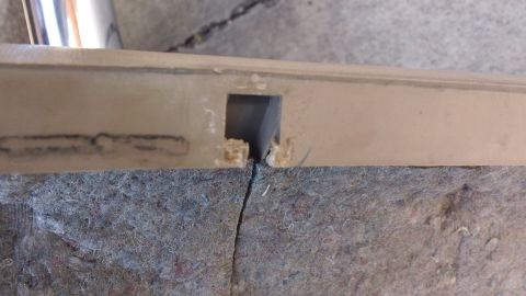 1994 Ford Bronco - Close-up of a tan door panel edge laid face-down on gray jute backing, showing a cracked and chipped cutout where a clip or switch mounts a…