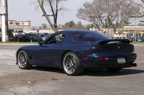 2006 Cadillac STS - Dark blue Mazda RX-7 (FD) coupe with aftermarket wheels and spoiler, seen from the rear 3/4 angle.