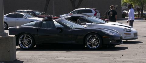 2006 Cadillac STS - A black Chevrolet Corvette C6 convertible is parked alongside a white Chevrolet Camaro, with other vehicles and people in the background.
