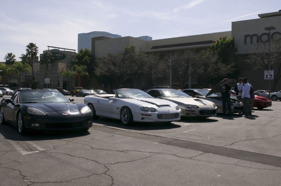 Several sports cars, including a black Corvette and a white convertible Camaro, parked in a lot with buildings in the background