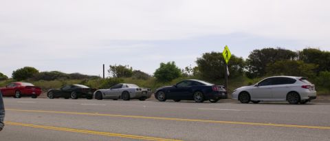 2006 Cadillac STS - A group of sports cars and a hatchback are parked side-by-side on the shoulder of a road with vegetation in the background.