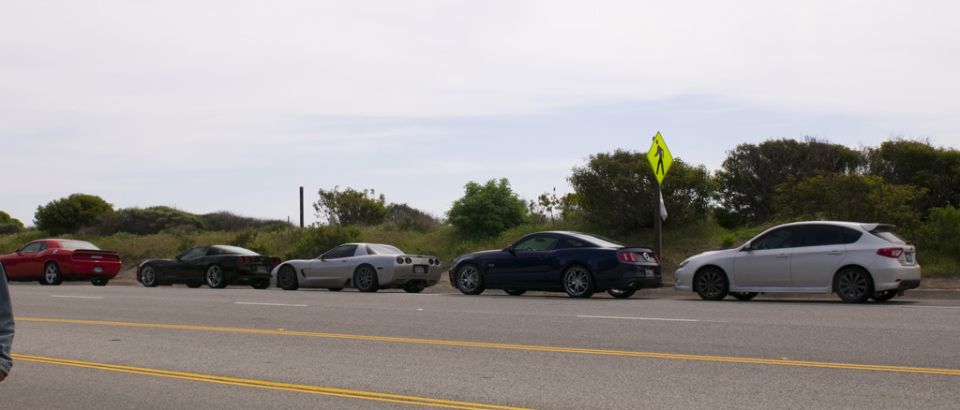 Several cars, including a red Dodge Challenger, two Chevrolet Corvettes, a Ford Mustang, and a Subaru Impreza, parked in a line along a roadside