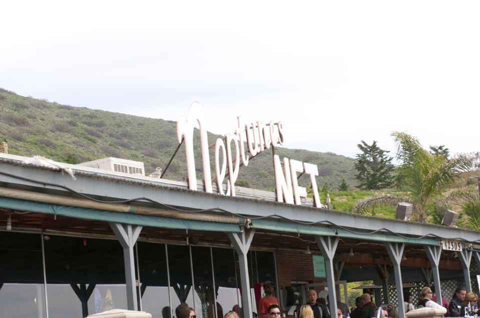 Building with a white sign that reads "Neptunes NET" against a cloudy sky and green hills
