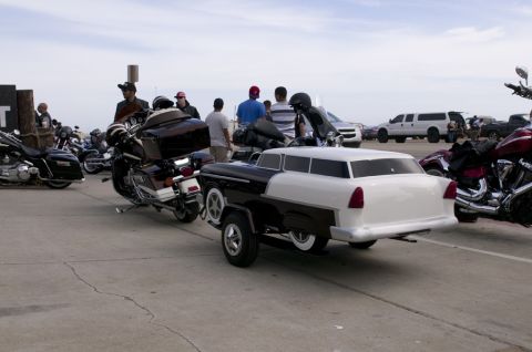 2006 Cadillac STS - A trailer designed as a vintage car is parked among motorcycles and other vehicles under a cloudy sky.