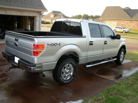 1995 Ford Bronco - Three-quarter rear view of a silver four-door crew cab Ford F-150 pickup parked on a wet residential driveway in front of an open garage. I…
