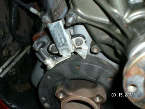 1996 Ford Bronco - Close-up of an engine front cover area with a metal sensor bracket, hardware and crankshaft hub flange.