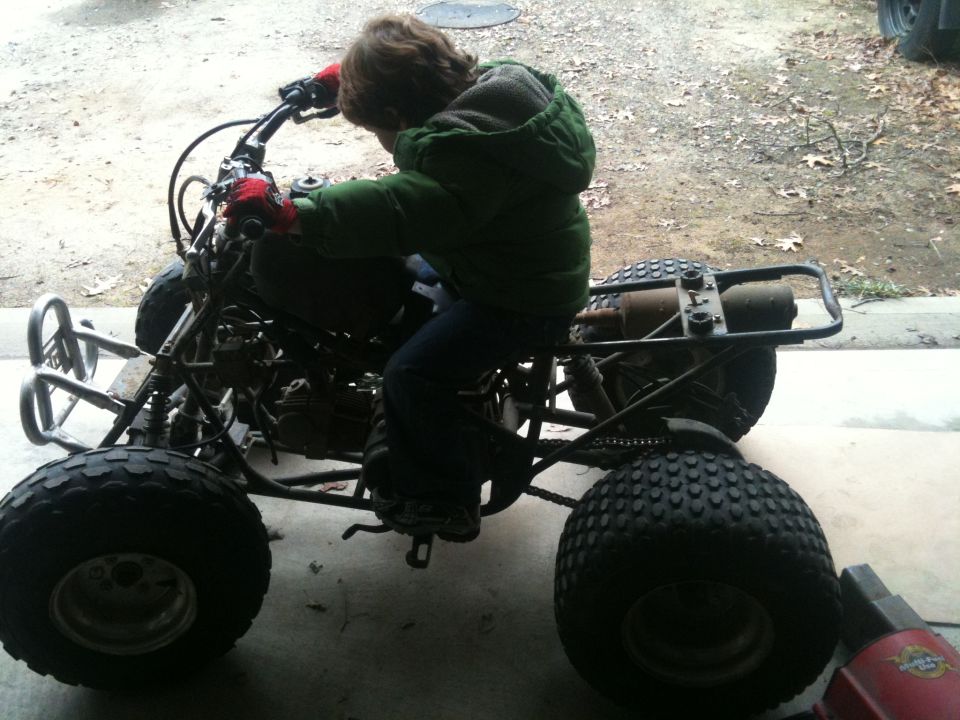 Child sitting on a small stripped-down ATV with bare frame and exposed engine in a garage