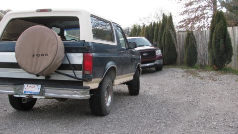 1992 Ford Bronco - A Ford Bronco and a Ford F-150 are parked in a gravel driveway next to a wooden fence and cypress trees.