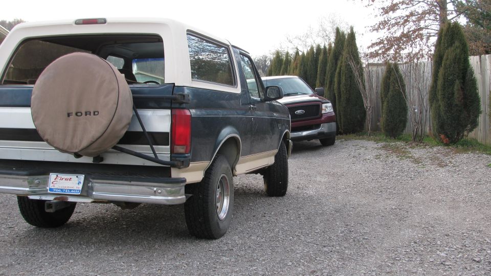 Rear view of a dark blue and tan Ford Bronco with a spare tire on the back, parked next to a red Ford F-150