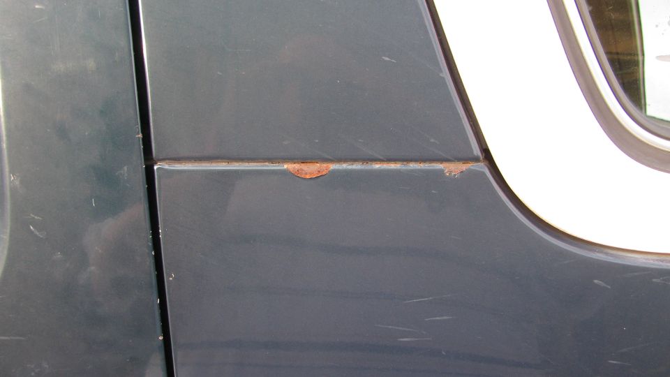Close-up of rust and paint blistering along a seam on a dark gray vehicle body panel