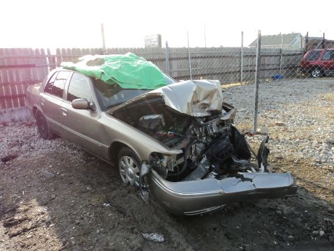 2004 Mercury Grand Marquis - This gold sedan sustained heavy front-end damage, with the hood and engine exposed and a tarp covering the roof.