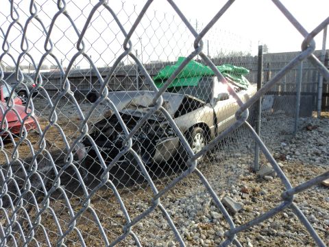 2004 Mercury Grand Marquis - A damaged sedan sits behind a fence, partially covered by a green tarp, with another vehicle visible in the background.