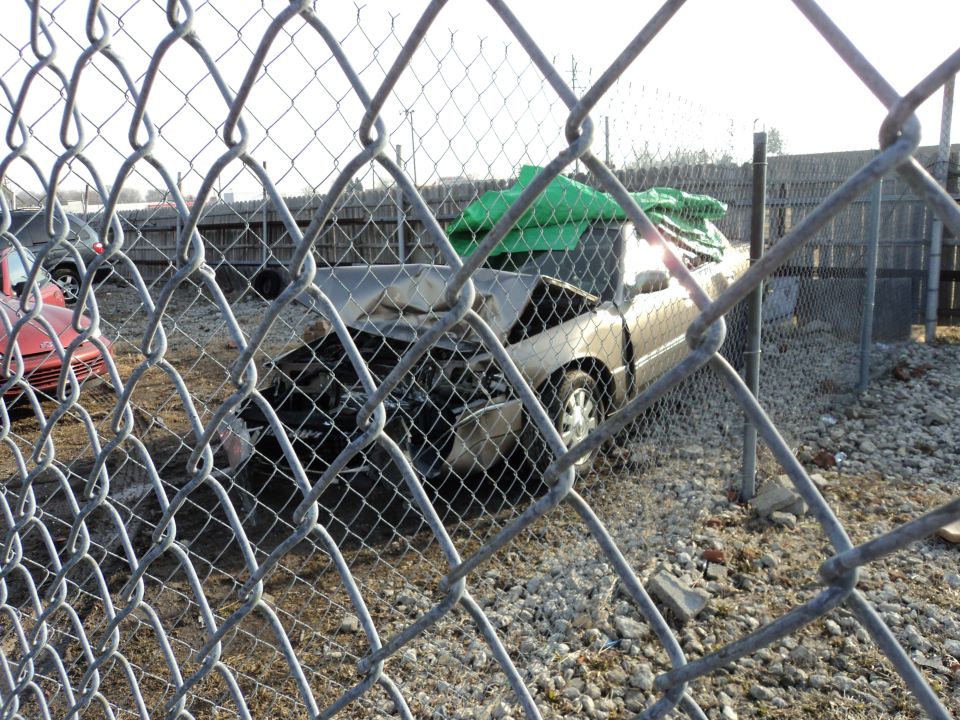 Damaged gold sedan behind a chain-link fence, engine exposed, green tarp on roof. Red car visible to the left