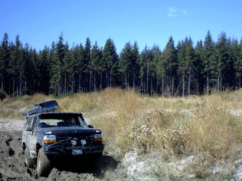 1995 Ford Bronco - A black Ford Explorer navigates a muddy trail with another vehicle behind it, surrounded by dry grass and a forest.