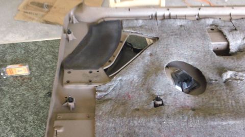 1994 Ford Bronco - A removed tan molded carpet set laid upside down on a shop floor, showing the jute backing, cut holes for the seat belt anchors and shifter…